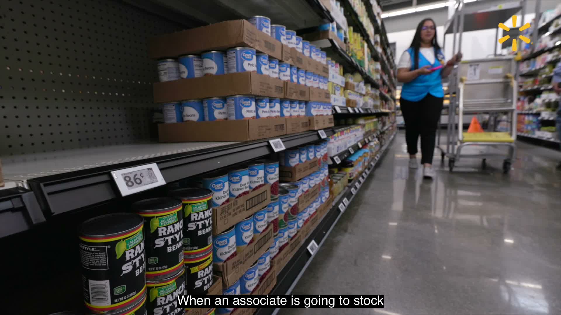 A Walmart associate uses a handheld device beside a replenishment cart and shelves of canned products.