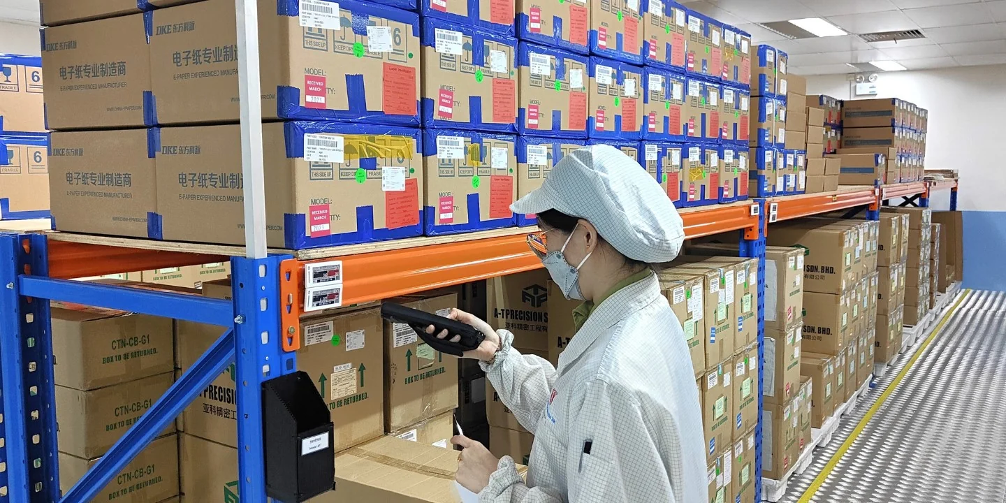 A PTSN warehouse operator uses a handheld PDA beside electronic inventory labels on a material rack.